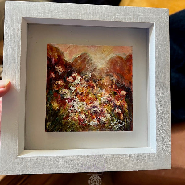 Framed miniature oil painting of sunlit flowers in a meadow 
with a white frame