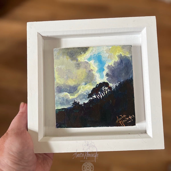 Small landscape painting in a white frame of trees against a bright cloudy sky
