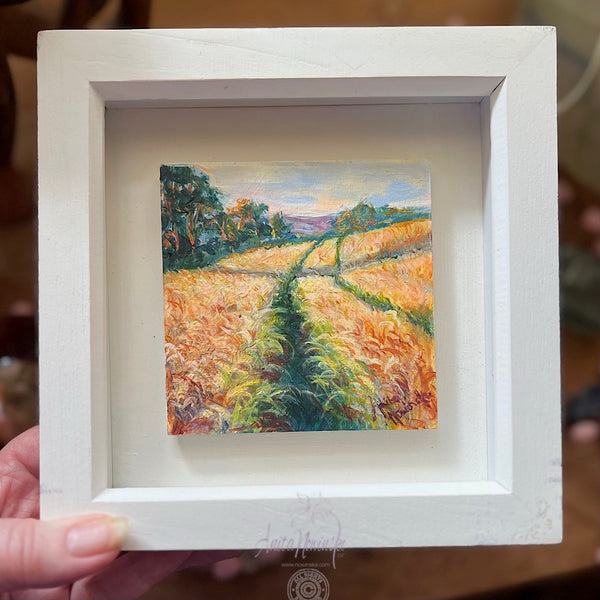 Miniature oil painting of a landscape of wheat fields at golden hour with paths and trees in a white frame .